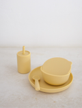 Rommer Co's Dinner Set is a minimalist collection of beige dishware, ideal as a baby gift. This set includes a lidded bowl, a cup with a straw, and a spoon. Crafted from premium silicone, the items are non-toxic and feature smooth surfaces and simple designs against an off-white background.