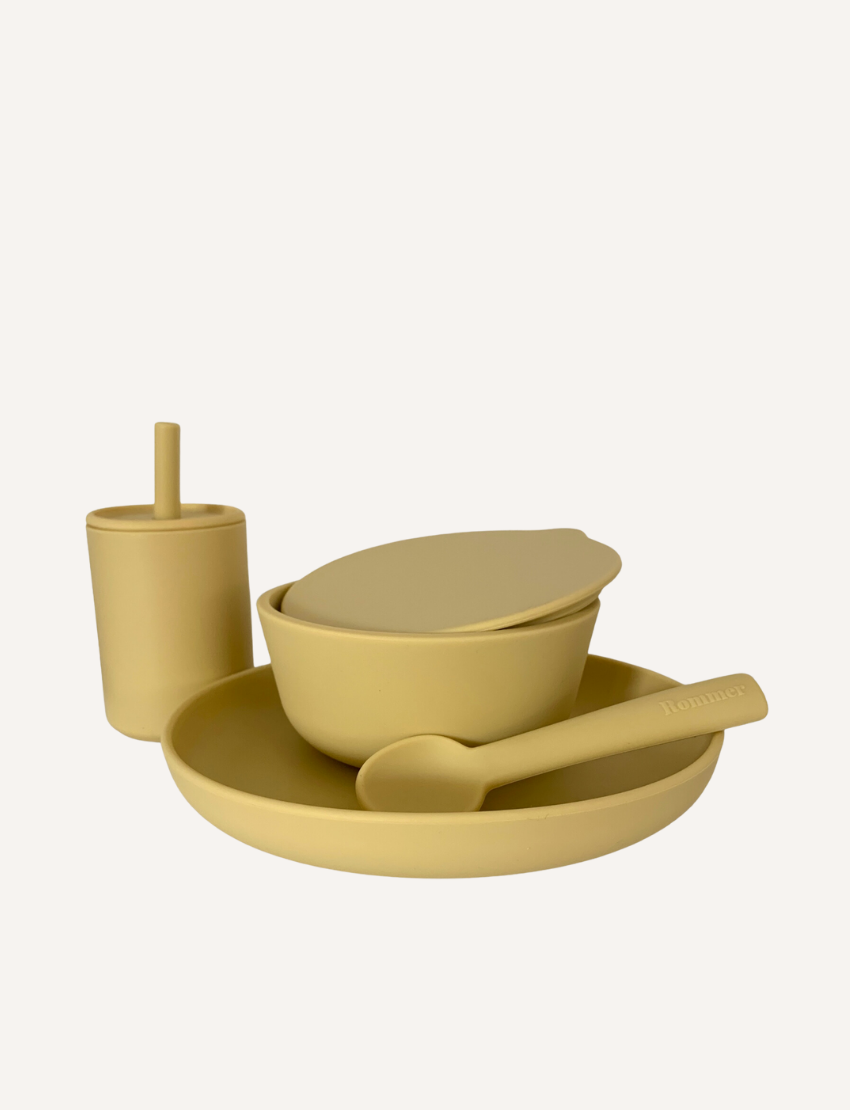 The Rommer Co Dinner Set is a peach-colored premium silicone set for children, featuring a cup with a straw, bowl with lid, plate, and spoon. Non-toxic and safe for the dishwasher and microwave, all items are neatly displayed against a plain white background.