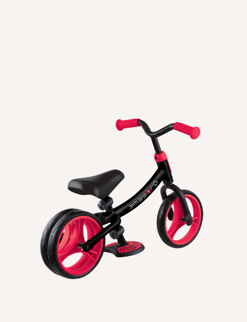 The Globber Go Bike Duo Balance Bike is a small, black and red toddler bike with no pedals, featuring EVA foam wheels. It has a black frame, a red adjustable handlebar, a black seat, and red wheels. The bike also includes a small footrest platform on the frame.