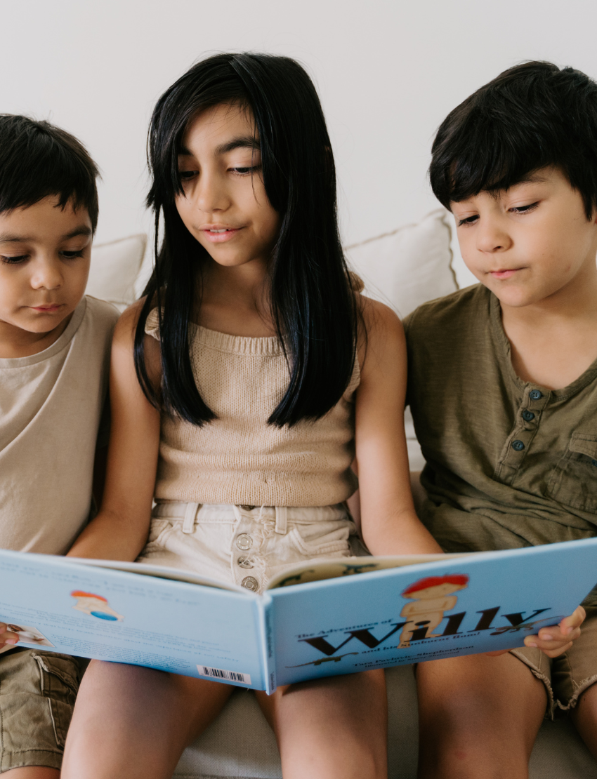 Three children are sitting on a couch, engaging with their Sensory Friendly Sunscreen Kit by The Play Way. The child in the middle is holding the kit, while the other two children are eagerly looking at it. All three appear focused on exploring the contents of the kit.