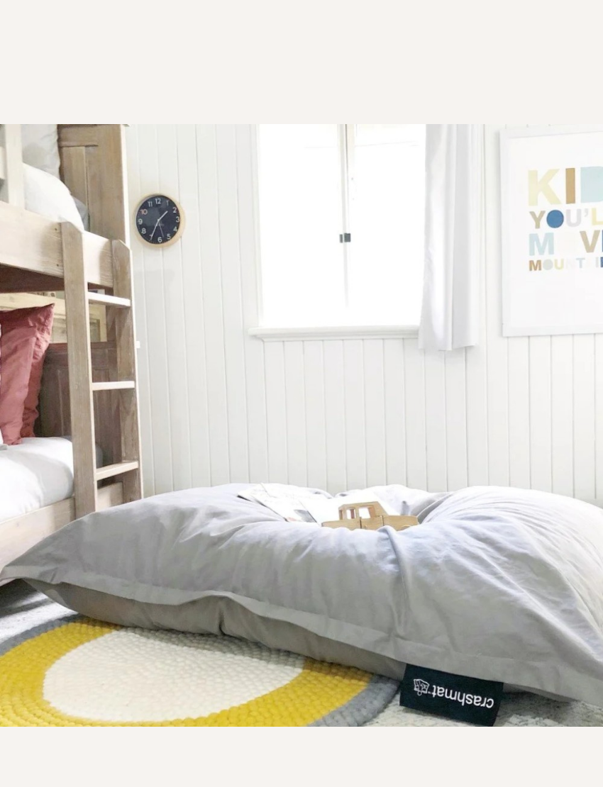 A cozy room showcases a wooden bunk bed, a large Crashmats Crashmat Beanbag in light grey on the floor, and a colorful circular rug. A wooden toy rests on the bean bag. A clock hangs on a white paneled wall near a window with curtains, complemented by a framed poster, creating a child-safe haven.