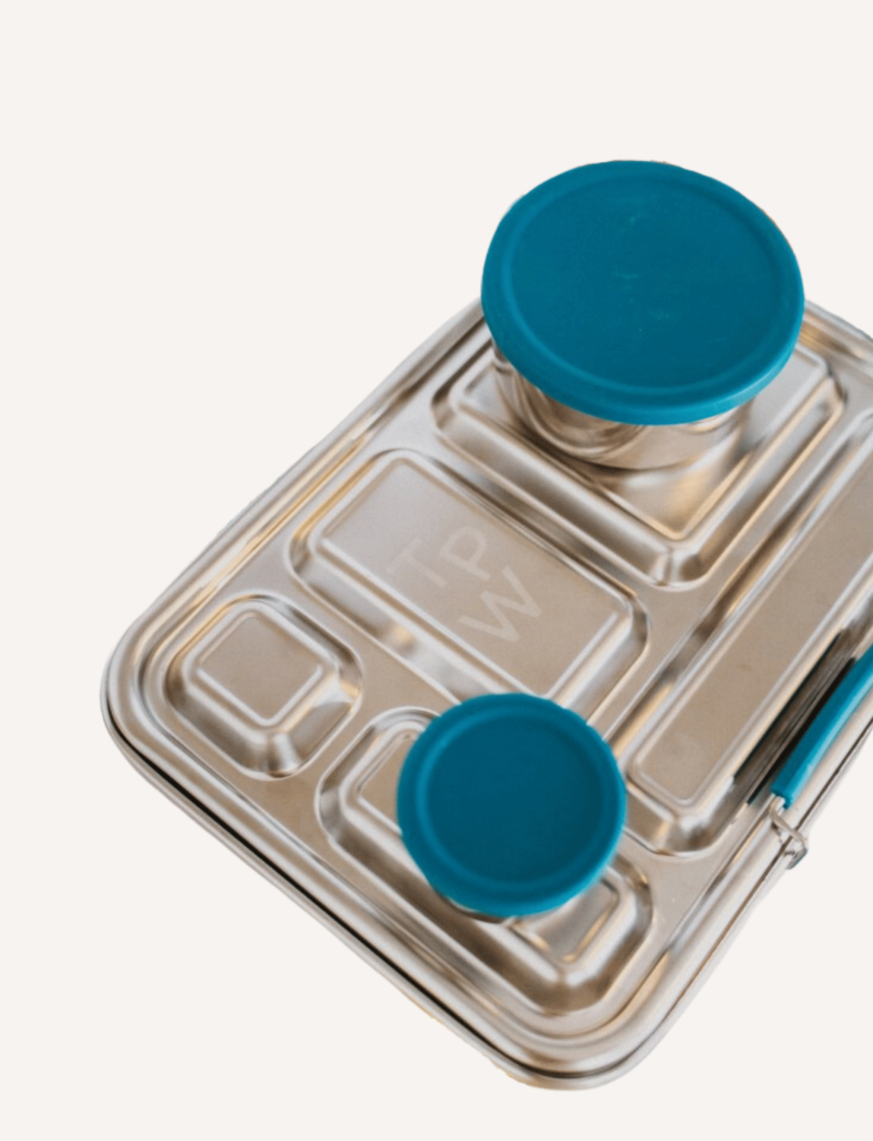 A child is seated at a table, reaching into The Play Way’s Lunch Buddy Bento Set filled with various foods, including fresh fruits and vegetables. The stainless steel bento set is placed on a wooden surface, and there are additional containers and some cucumbers nearby.