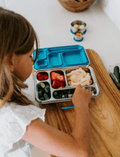 A child is seated at a table, reaching into The Play Way’s Lunch Buddy Bento Set filled with various foods, including fresh fruits and vegetables. The stainless steel bento set is placed on a wooden surface, and there are additional containers and some cucumbers nearby.