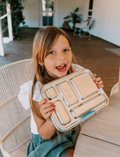 A child is seated at a table, reaching into The Play Way’s Lunch Buddy Bento Set filled with various foods, including fresh fruits and vegetables. The stainless steel bento set is placed on a wooden surface, and there are additional containers and some cucumbers nearby.