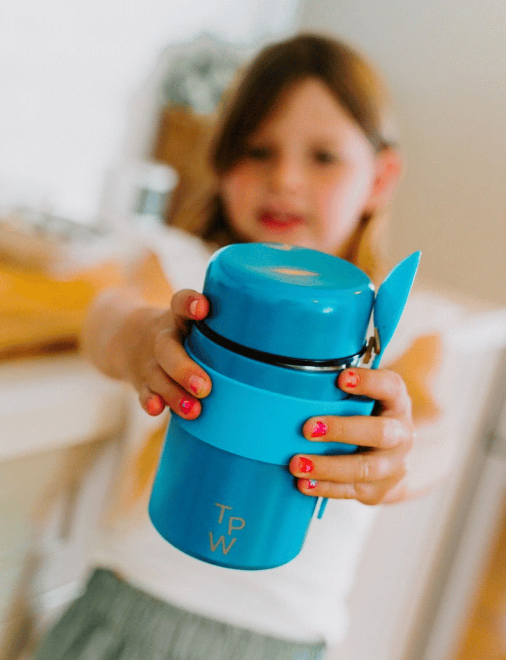 A young girl holding a small blue Lunch Buddy Insulated Container from The Play Way, featuring a leak-proof lid and the brand's initials "T P W" engraved on it. The focus is on the stainless steel container, with the girl slightly blurred in the background. Her nails are painted pink.