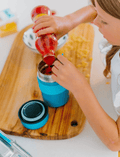 A young girl holding a small blue Lunch Buddy Insulated Container from The Play Way, featuring a leak-proof lid and the brand's initials 