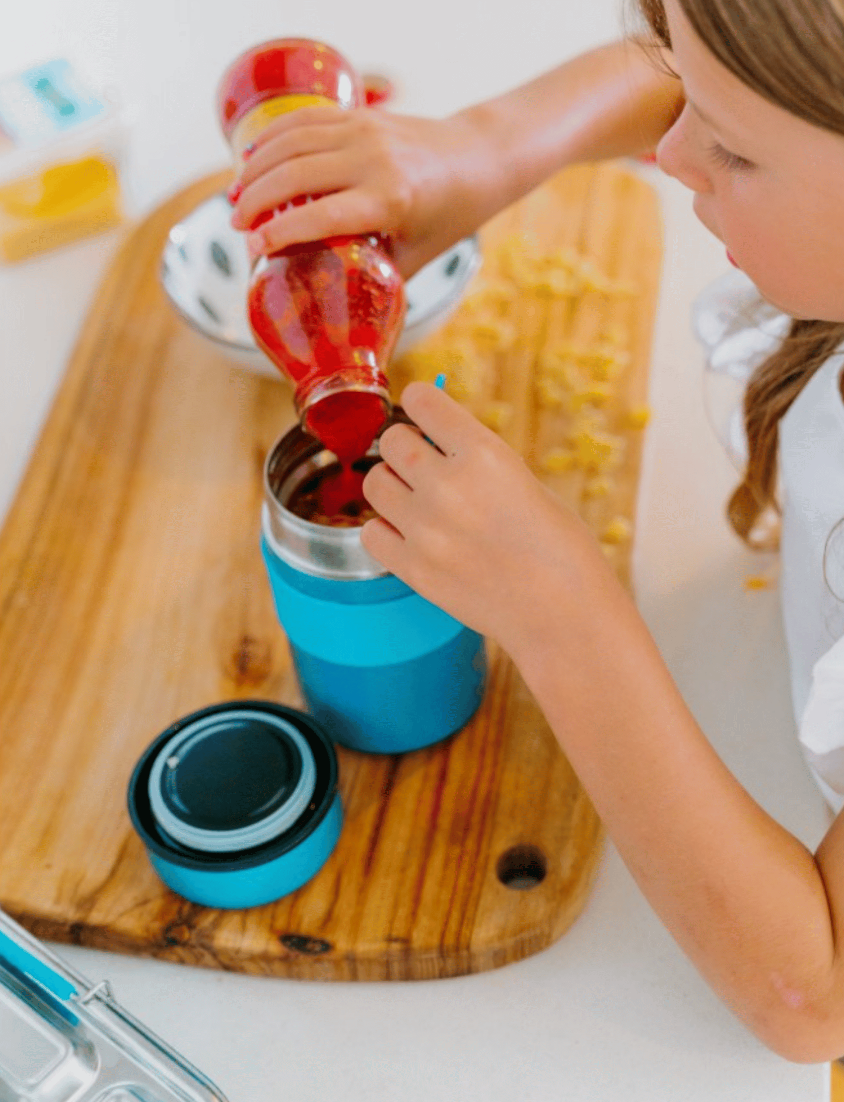 A young girl holding a small blue Lunch Buddy Insulated Container from The Play Way, featuring a leak-proof lid and the brand's initials "T P W" engraved on it. The focus is on the stainless steel container, with the girl slightly blurred in the background. Her nails are painted pink.