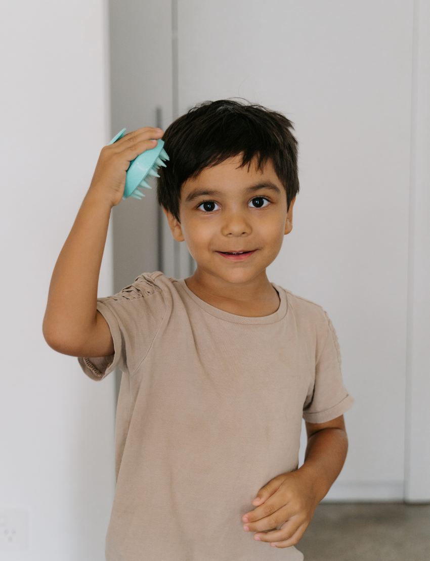 A turquoise Melo Hair Care Kit scalp therapy massager by The Play Way, featuring a round design and soft bristles, is pictured next to its blue box packaging. The box includes text instructions and caution warnings. The background is white.