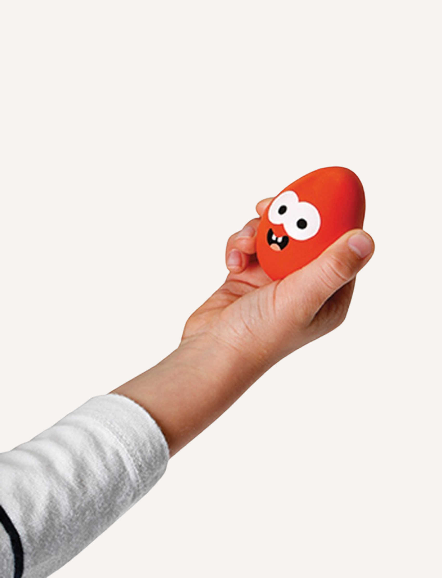 A hand is holding a red egg from the Janod Egg and Spoon Race, featuring cartoonish white eyes and a small smiling mouth, reminiscent of a children's game. The background is plain white. The sleeve of the arm holding the egg appears to be white with black cuffs, contrasting nicely against the playful toy.