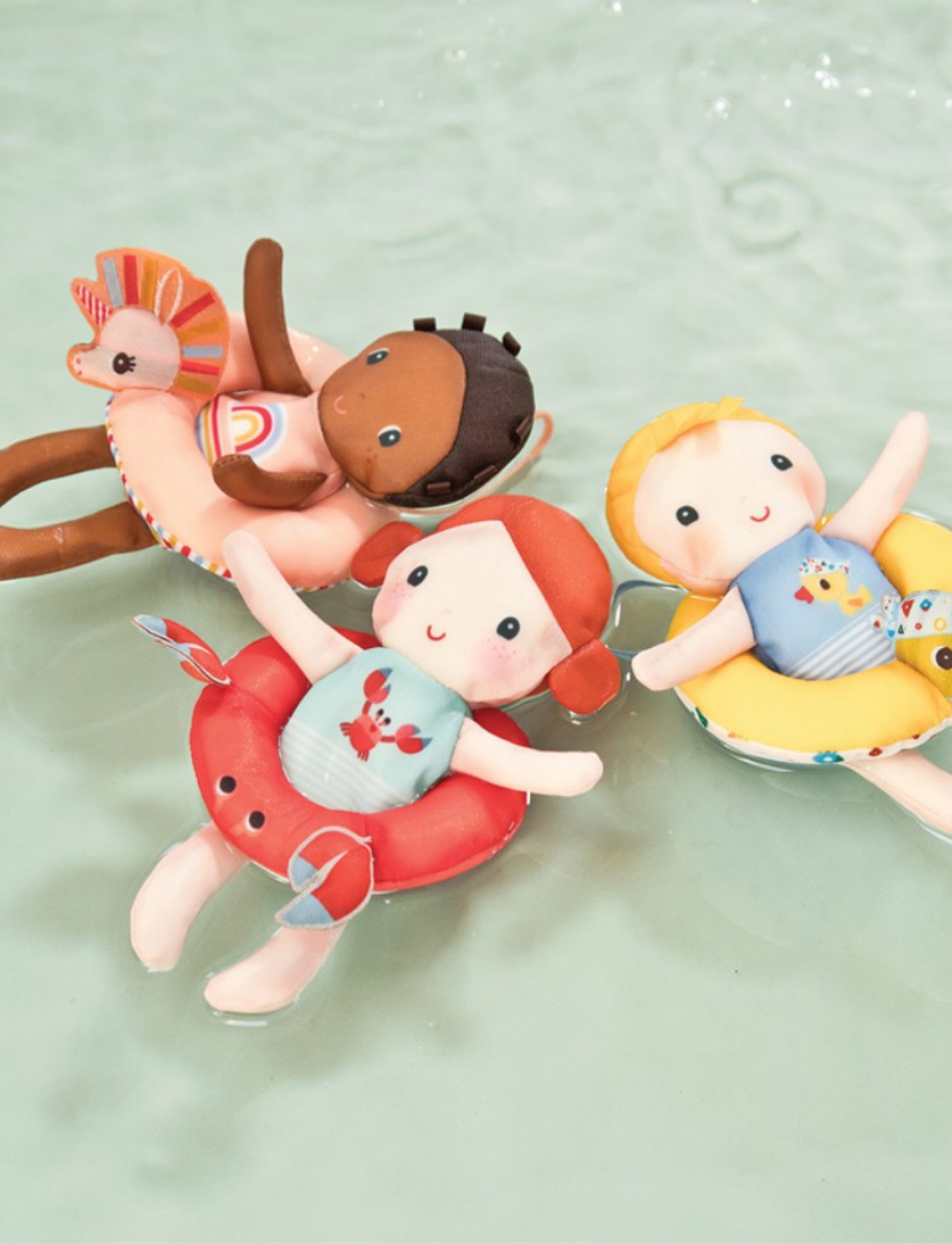 Two plush toys are shown against a plain background. One is a soft bath doll named Lena with dark brown skin, wearing an outfit with a rainbow design from the Lilliputiens brand's Bath Doll - Unicorn collection. The other is a pink circular ring toy resembling a unicorn floatie, complete with a small rainbow attachment and a loop handle at the top.