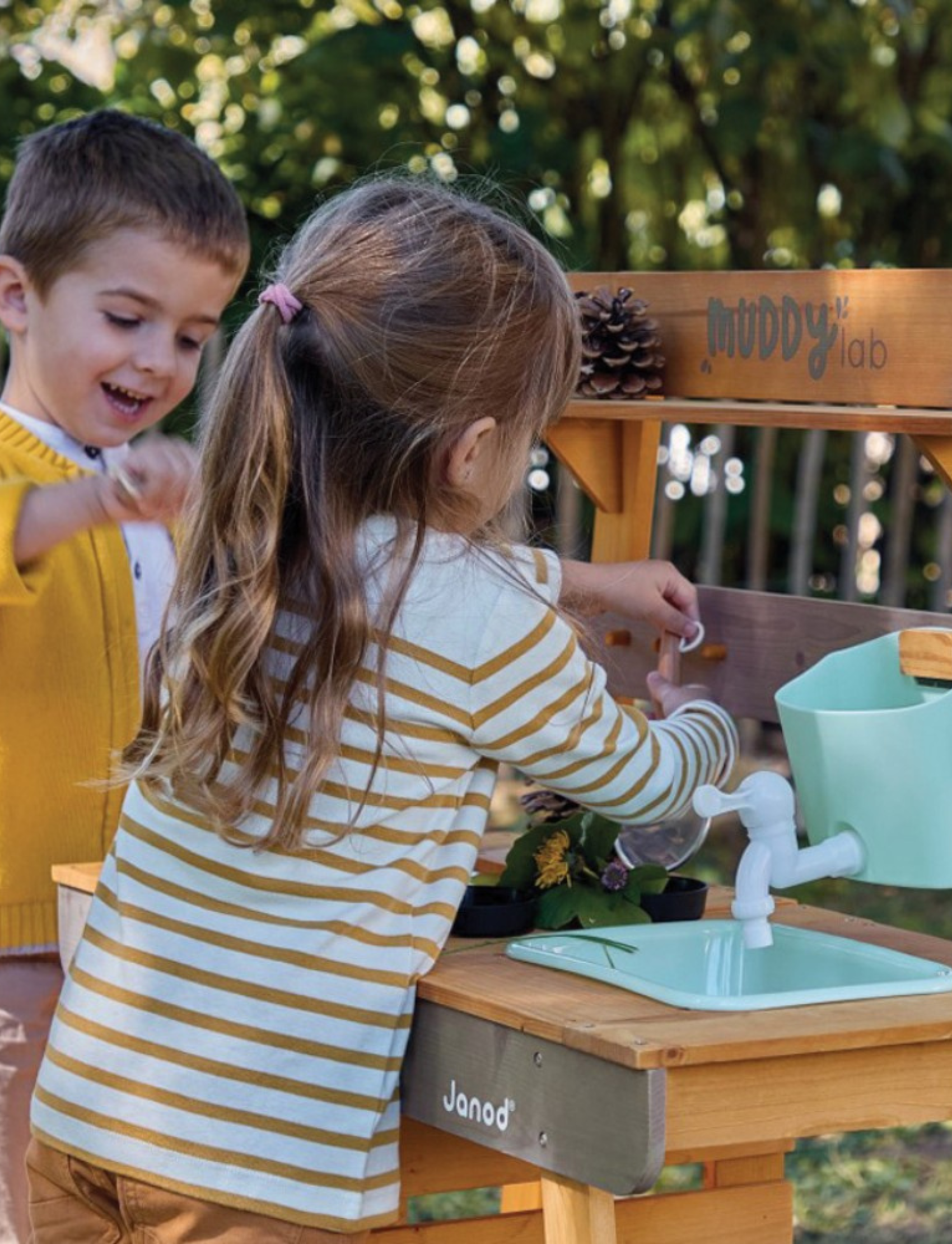 Janod's "Muddy Lab Outdoor Kitchen" is a boxed toy set that includes an outdoor wooden kitchen crafted from FSC certified wood. It features shelves, hooks for utensils, and an attached water dispenser. The packaging displays images of the assembled product alongside children joyfully playing with the Muddy Lab Outdoor Kitchen.