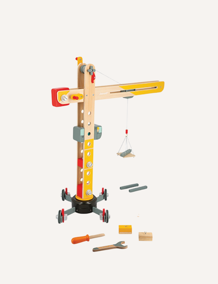 The image features the Janod Giant Crane, a wooden toy that resembles a large construction crane with a base, movable arm, and pulley system. Scattered around its base are various small tools and accessories, including a screwdriver and wrench, making it a delightful role-play toy.