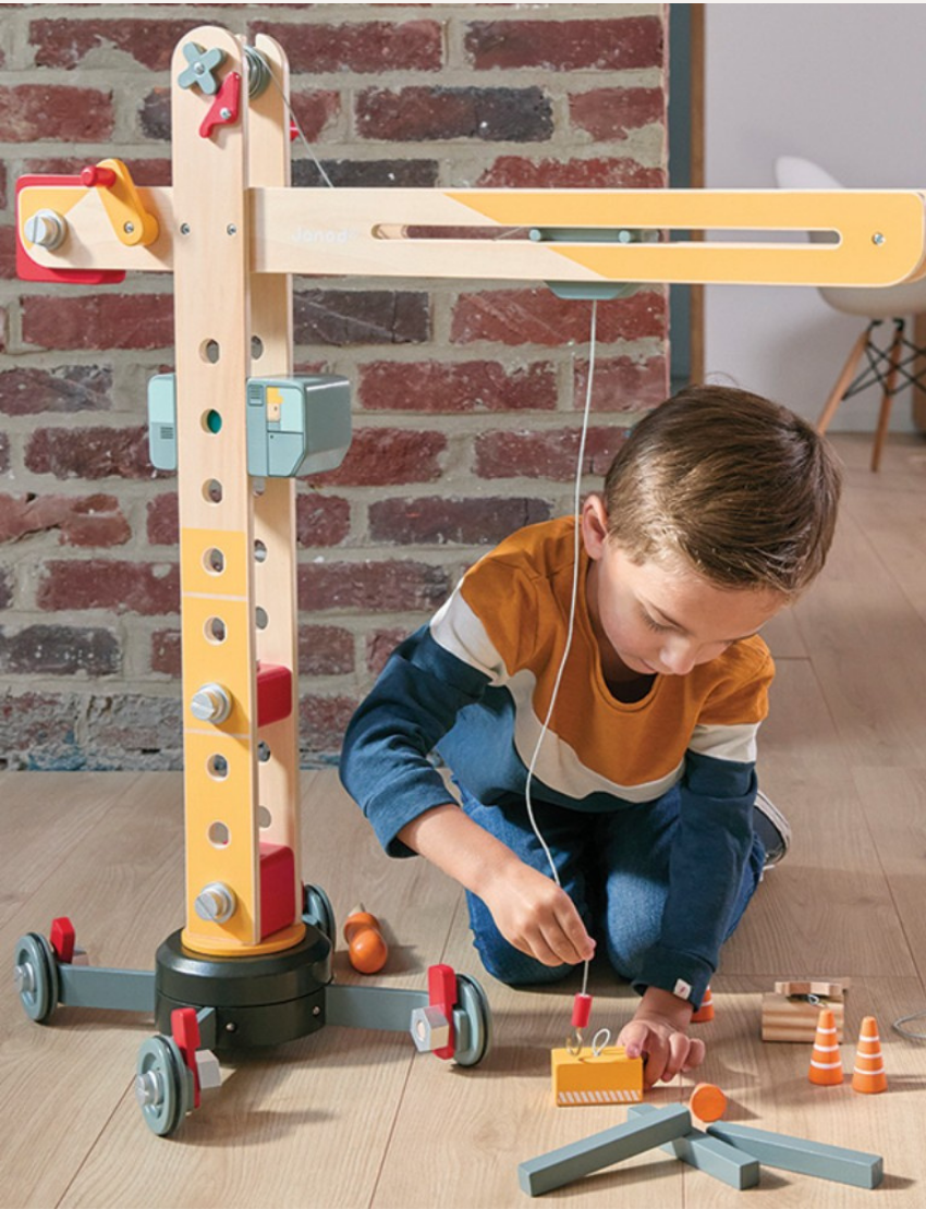 The image features the Janod Giant Crane, a wooden toy that resembles a large construction crane with a base, movable arm, and pulley system. Scattered around its base are various small tools and accessories, including a screwdriver and wrench, making it a delightful role-play toy.