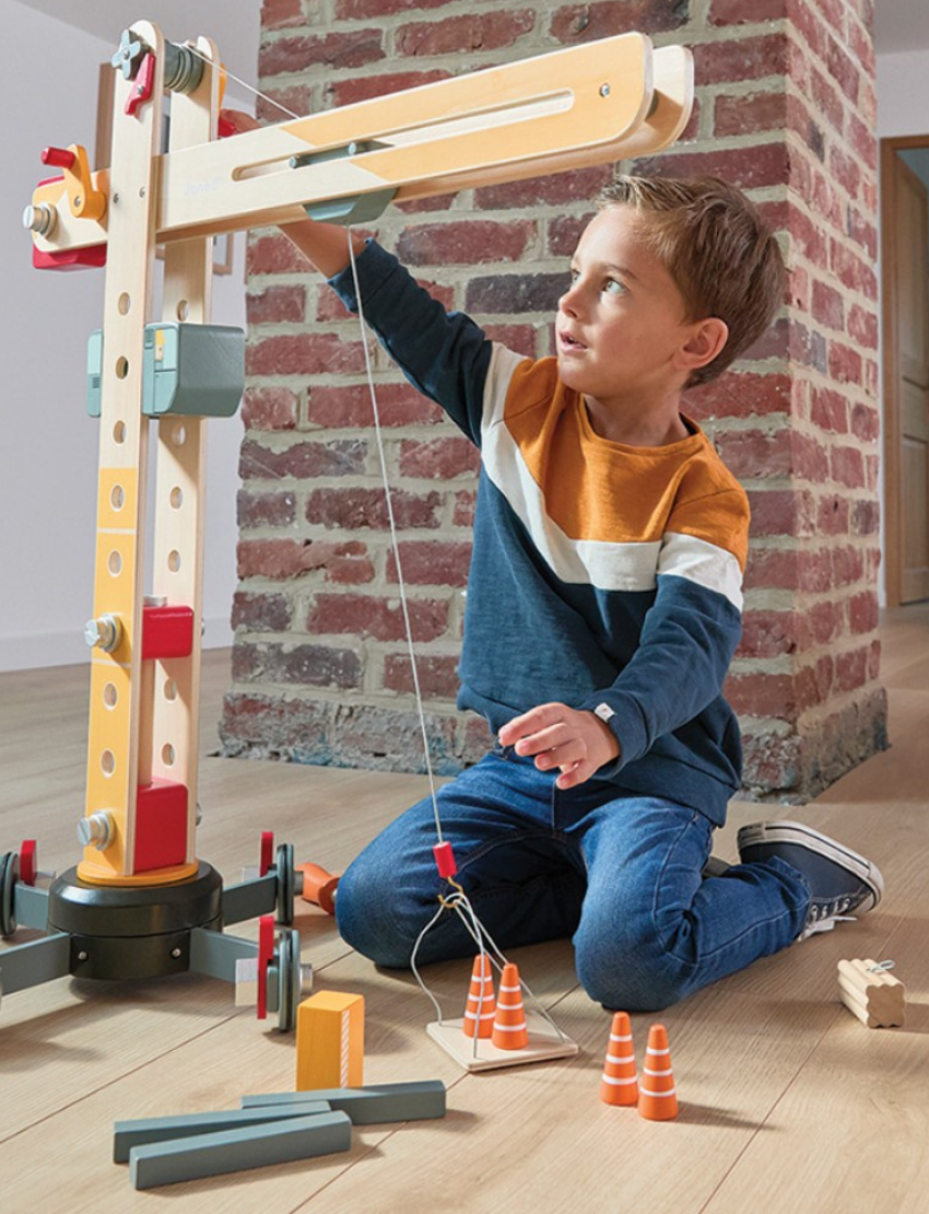 The image features the Janod Giant Crane, a wooden toy that resembles a large construction crane with a base, movable arm, and pulley system. Scattered around its base are various small tools and accessories, including a screwdriver and wrench, making it a delightful role-play toy.