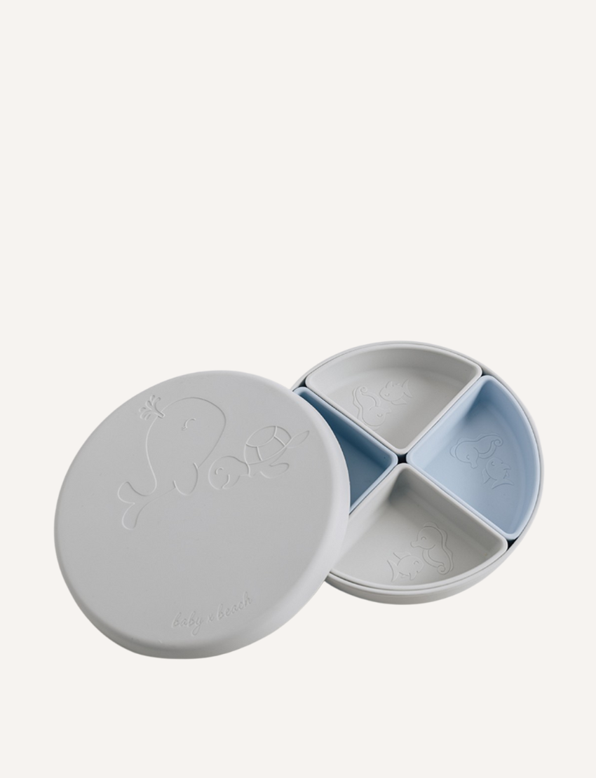 The "Multi-use Silicone Divider Plate" by Baby By Beach is a stylish, multifunctional round baby plate with a lid and three sections. Made from food-safe silicone, it features light blue and off-white compartments adorned with embossed whale and fish designs for practicality.