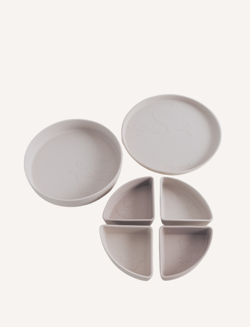 The "Multi-use Silicone Divider Plate" by Baby By Beach is a stylish, multifunctional round baby plate with a lid and three sections. Made from food-safe silicone, it features light blue and off-white compartments adorned with embossed whale and fish designs for practicality.