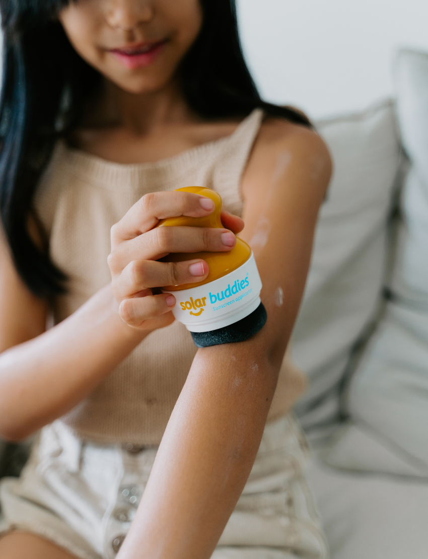 Three children are sitting on a couch, engaging with their Sensory Friendly Sunscreen Kit by The Play Way. The child in the middle is holding the kit, while the other two children are eagerly looking at it. All three appear focused on exploring the contents of the kit.