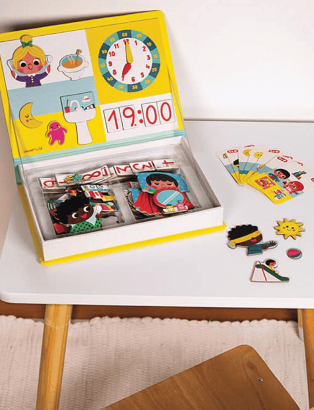 The "Learn Time Magnetic Book" by Janod is a children's educational toy set that includes a vibrant board with cartoon characters, a clock showing 12:15, and various magnetic pieces for storytelling and activity cards. The magnetic book is propped open, displaying all the pieces neatly within the storage area to help kids learn to tell the time.