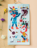 The Lilliputiens Pin The Tail On The Donkey children's game features characters in colorful costumes. The game board showcases vibrant cartoon animals and includes two separate cut-out costumes. A blue blindfold and extra costume pieces are conveniently placed next to the board.