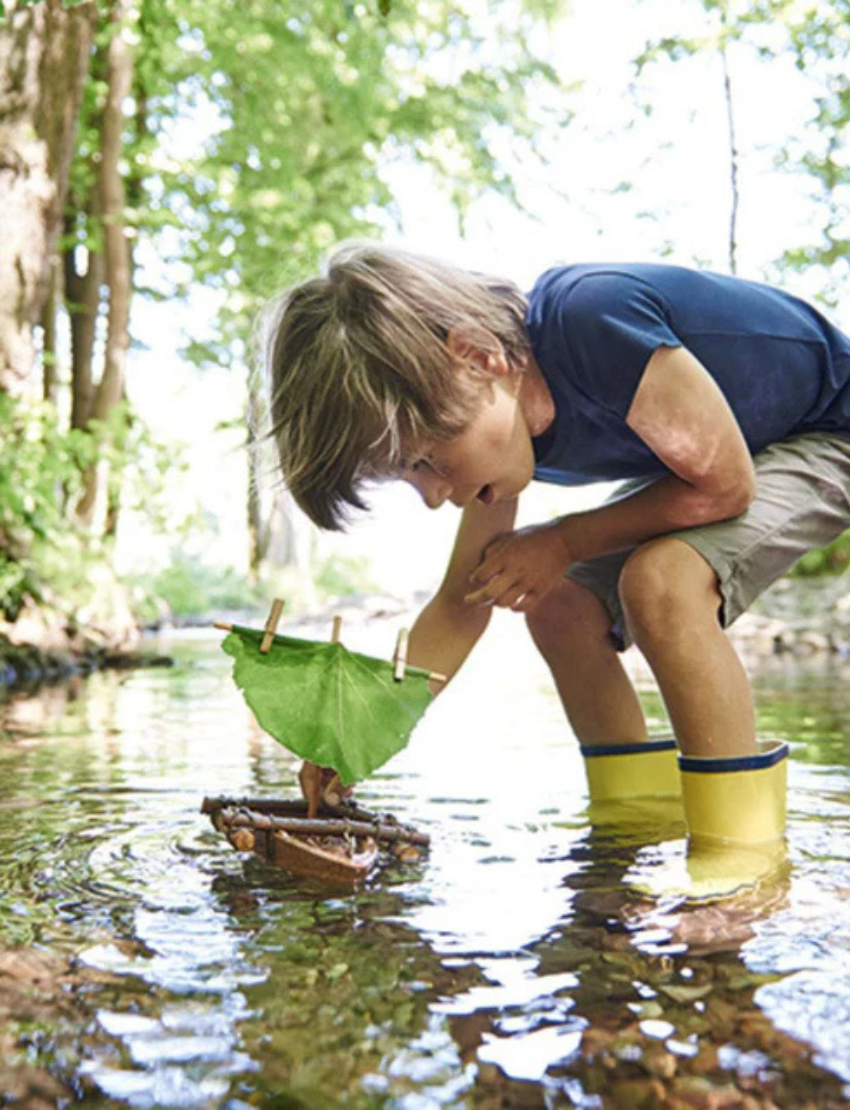 The HABA Cork Boat Kit includes a small model raft constructed from unsinkable cork and sticks, featuring an eco-friendly green leaf sail fastened with clothespins. It also comes with a piece of twine attached to the raft for easy maneuvering.