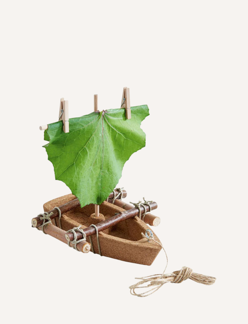 The HABA Cork Boat Kit includes a small model raft constructed from unsinkable cork and sticks, featuring an eco-friendly green leaf sail fastened with clothespins. It also comes with a piece of twine attached to the raft for easy maneuvering.