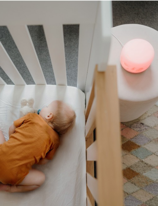 A baby wearing a brown onesie sleeps curled up in a white crib with wooden bars. A white plush toy rests next to the baby. Beside the crib, on a patterned rug, sits a small table featuring the Glow Dreaming Sleep Easy by Glow Dreaming, casting a soft pink light. The floor is carpeted.