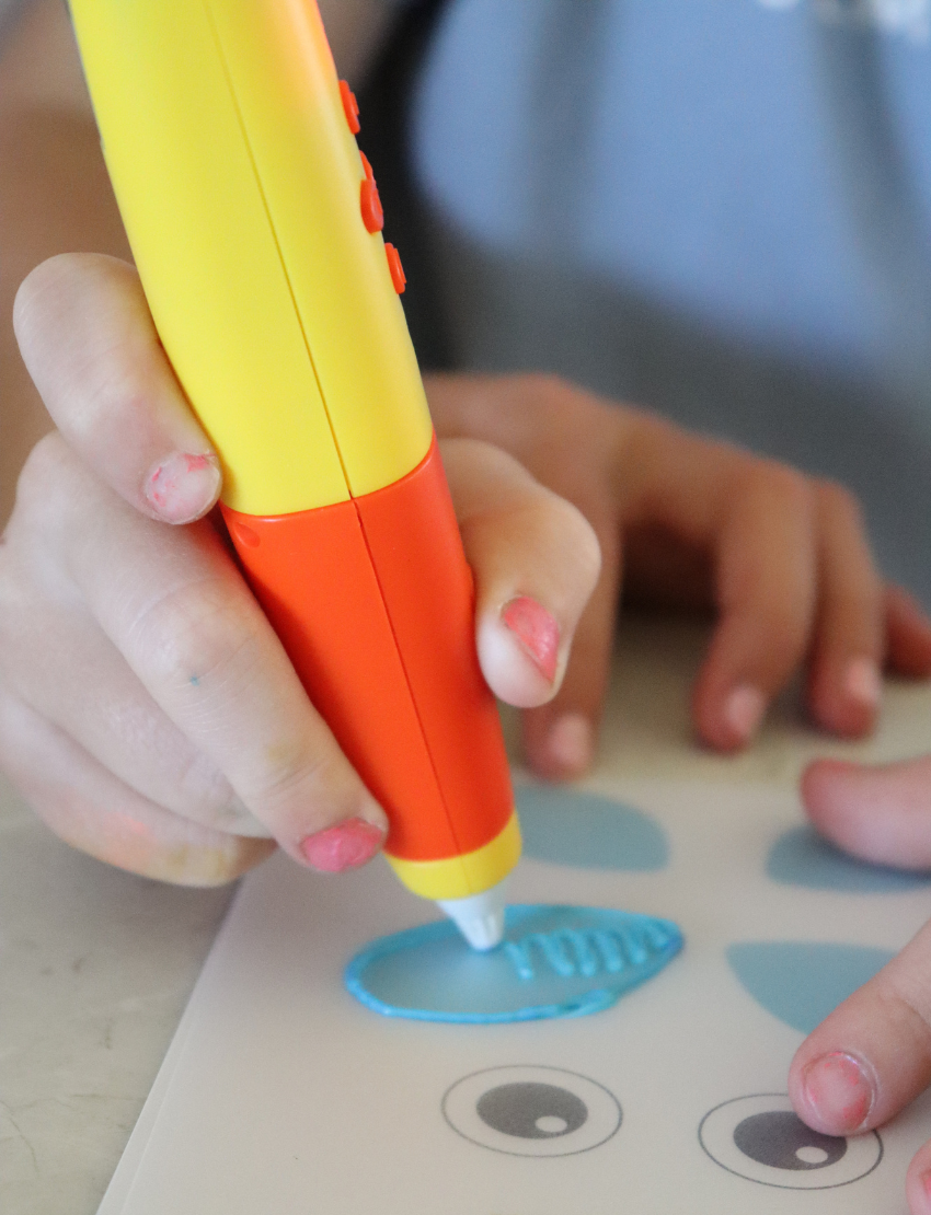 A packaging box for The Play Way's 3D Printing Pen is displayed. The box features images of the pen, 3D models of a house, a dinosaur, and the Eiffel Tower, with children in the background. Text on the box reads "3D Printing Pen" and provides some specifications.