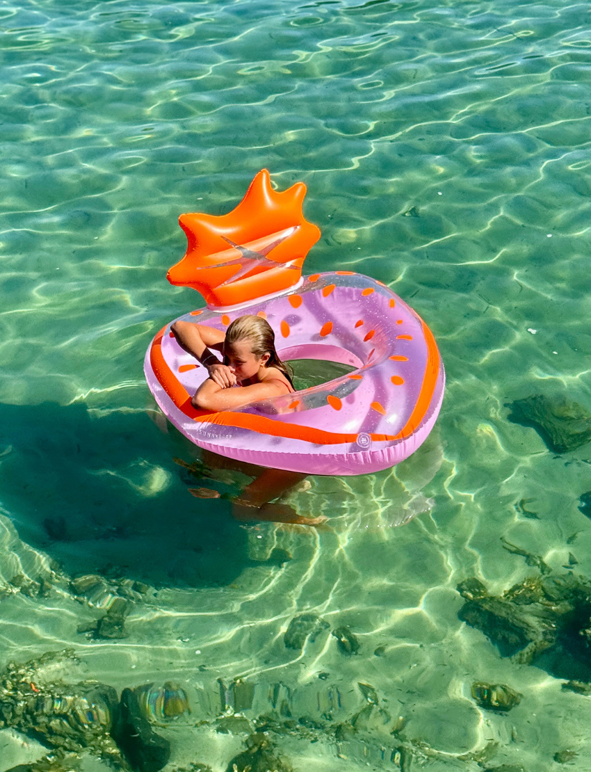 A black bird with a pointed beak is nestled inside a white mesh bag placed within a clear Tube Pool Ring on sandy ground. The float features the brand name "Sunnylife.