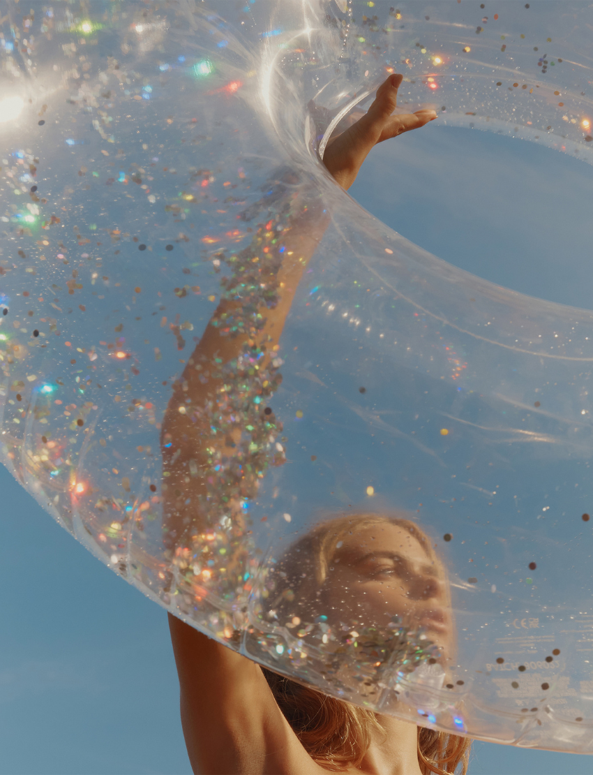 A black bird with a pointed beak is nestled inside a white mesh bag placed within a clear Tube Pool Ring on sandy ground. The float features the brand name "Sunnylife.