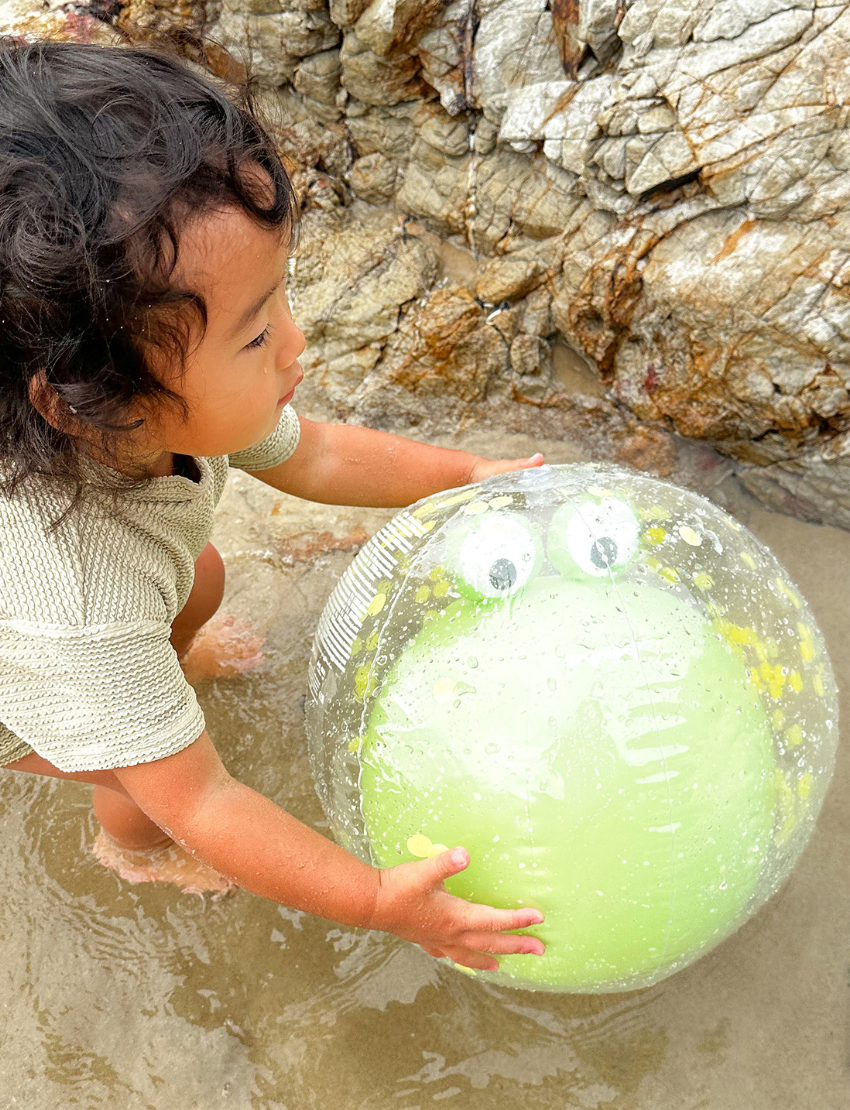 The image shows a clear plastic package displaying multilingual warning labels on its exterior. Inside, there is a partially visible aqua-colored 3D Inflatable Beach Ball from Sunnylife, along with a batch number and item code printed at the bottom.