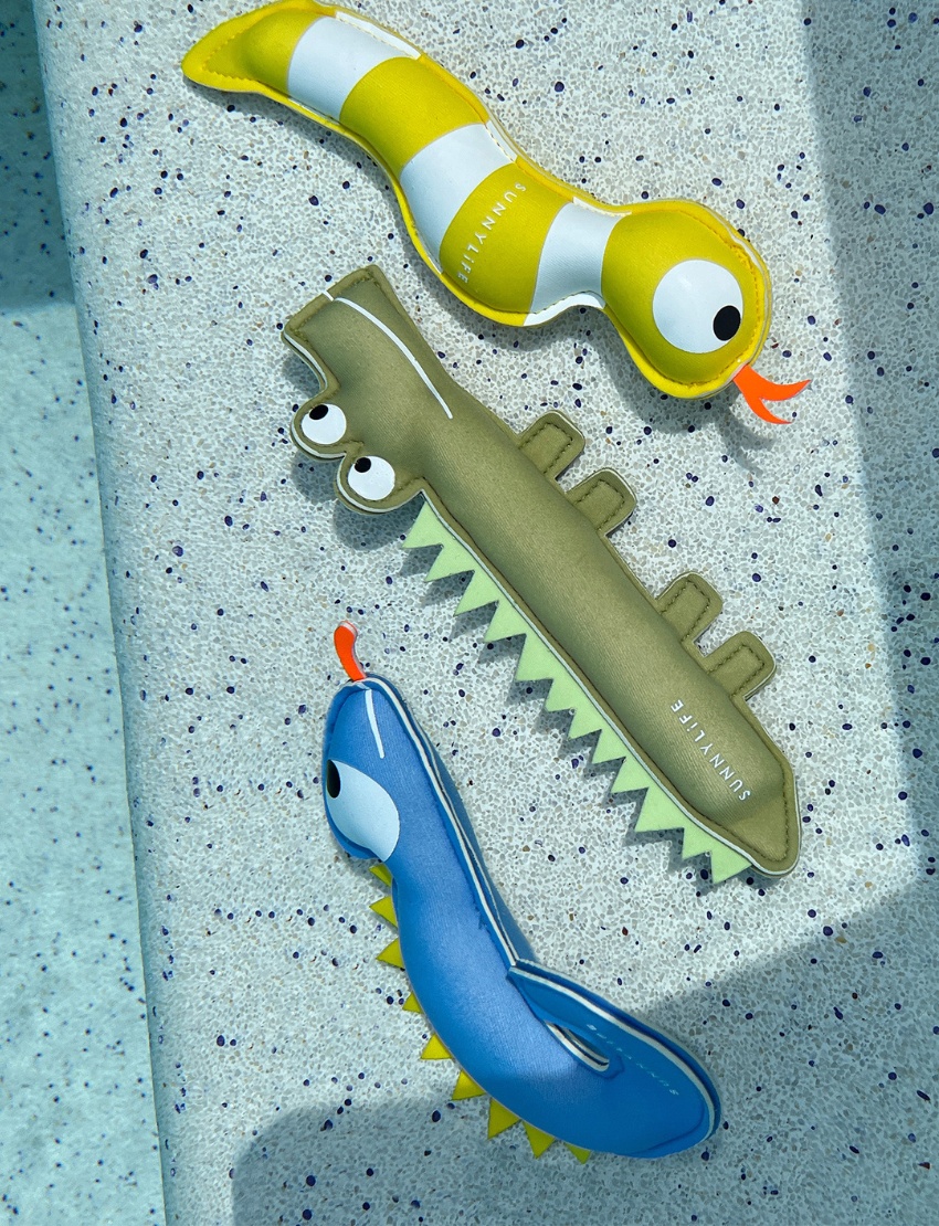 Three vibrant Dive Buddies from Sunnylife float on a swimming pool's surface. The toys feature a yellow and white striped design, a green spiky design, and a blue one with a red mouth. Sunlight gleams off the water, creating ripples and reflections.