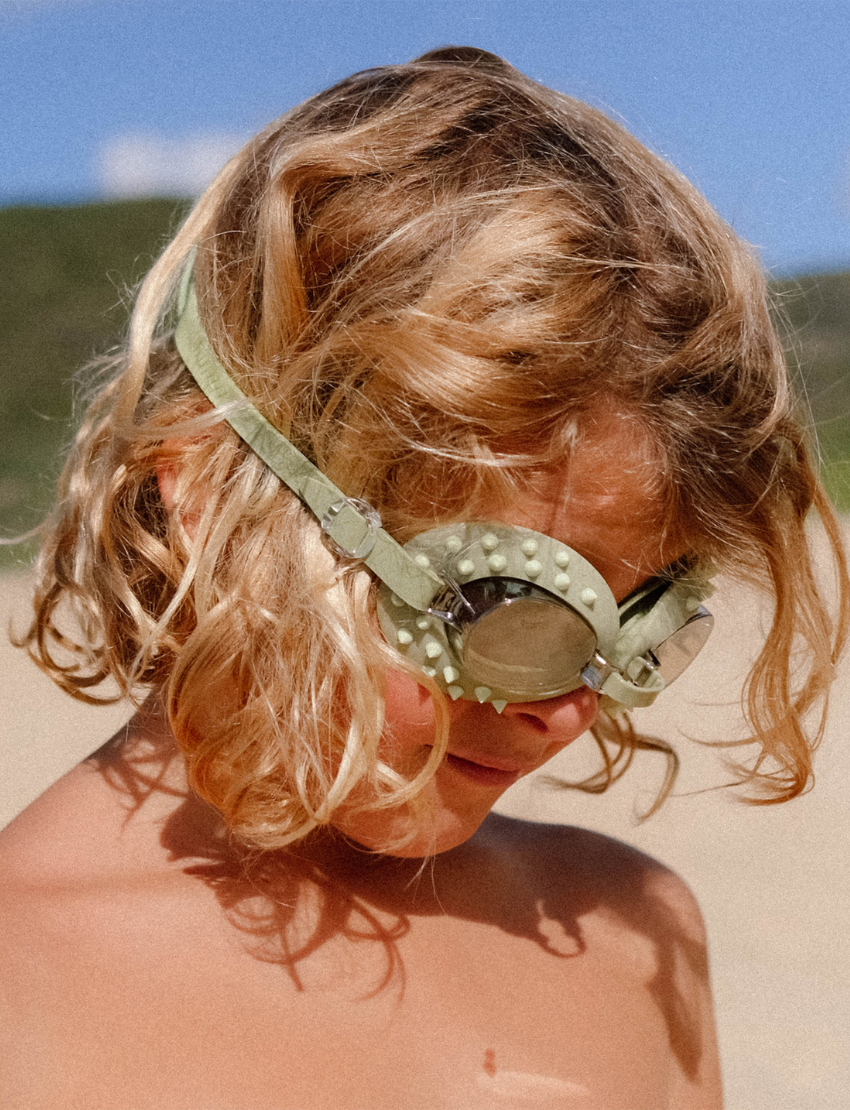 The Cookie The Croc Khaki Kids Swim Goggles from Sunnylife feature textured frames, an adjustable silicon strap, reflective anti-fog lenses for enhanced visibility, and unique small protrusions around the edges.