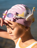 The Kids Swim Goggles by Sunnylife feature a playful swan-shaped head strap, with purple frames and reflective lenses. The head strap showcases a white body and yellow accents, complete with a crown. Both items are displayed against a plain white background.