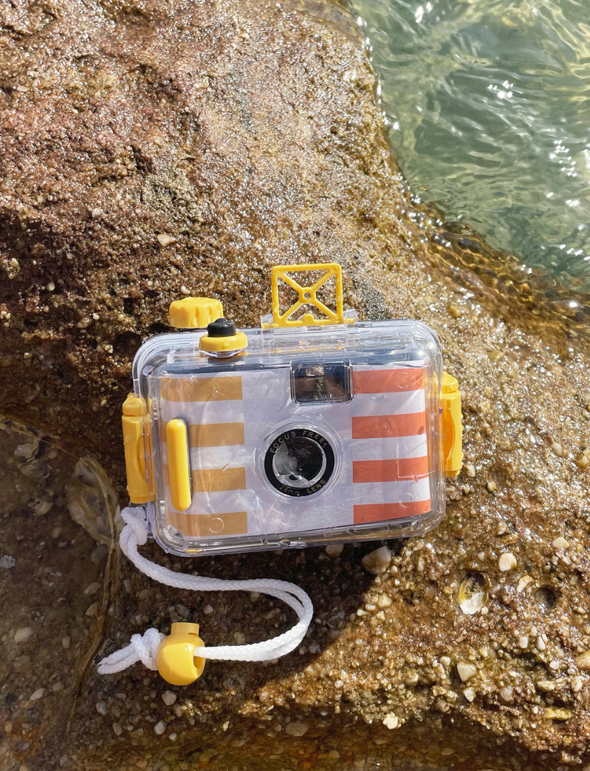 A Sunnylife Underwater Camera, encased in a transparent protective shell with yellow accents, rests on a textured stone surface. The camera boasts a focus-free 28mm lens and features a striped design in white, orange, and yellow.