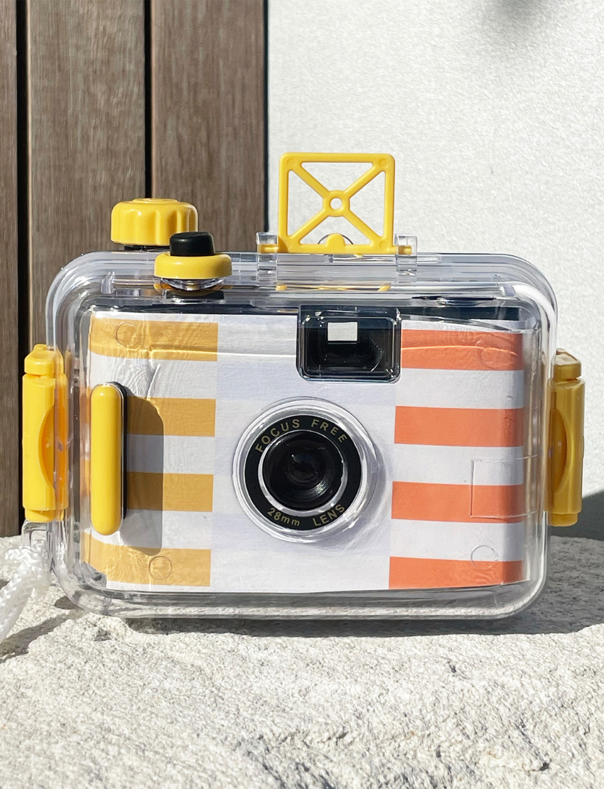 A Sunnylife Underwater Camera, encased in a transparent protective shell with yellow accents, rests on a textured stone surface. The camera boasts a focus-free 28mm lens and features a striped design in white, orange, and yellow.