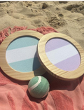 The Catch Ball Set - Rio Sun Multi by Sunnylife includes two round wooden paddles adorned with blue and lavender stripes, elegantly set against a light backdrop. A small green and white tennis ball floats above, inviting play.