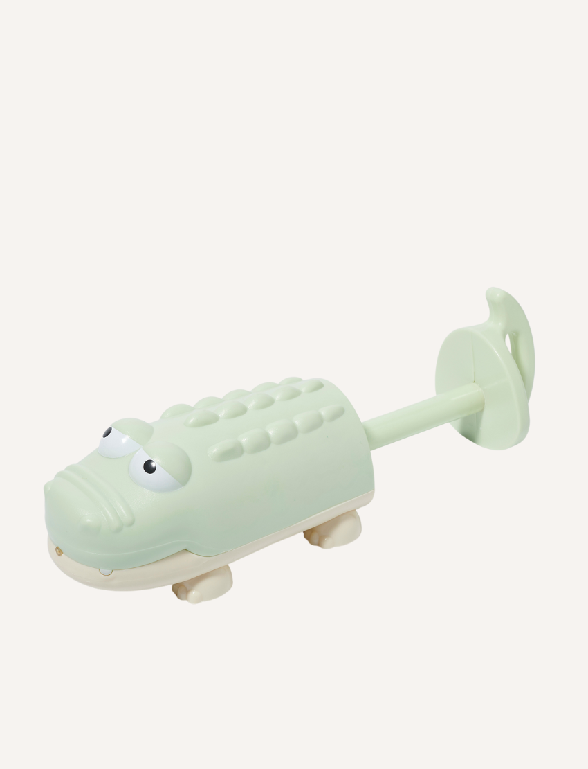 A close-up image of two ceramic water squirters on a marble surface. On the left is a purple dolphin with a smiling expression, and on the right is a light green alligator featuring cartoonish characteristics. The background is softly blurred. These are products from the Sunnylife brand.