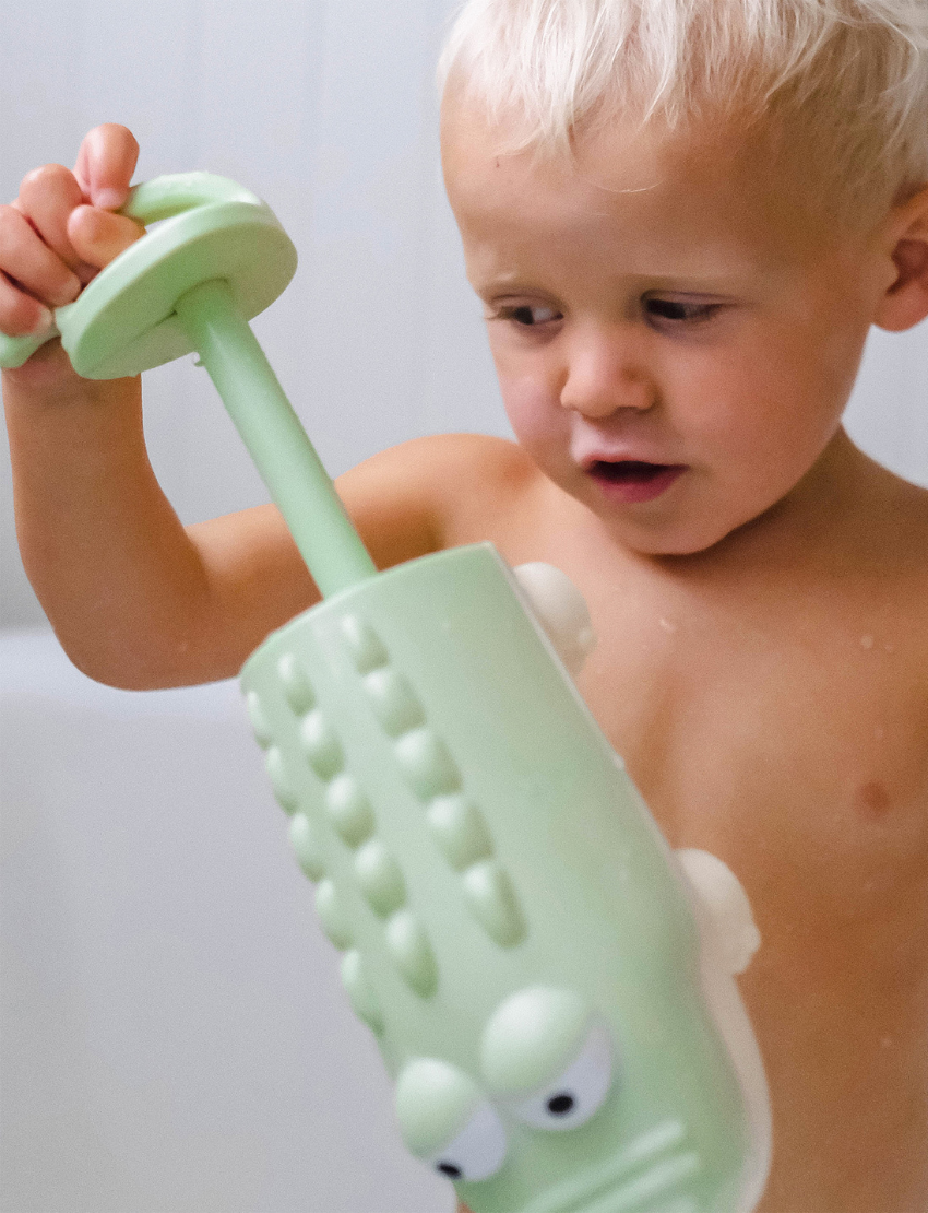 A close-up image of two ceramic water squirters on a marble surface. On the left is a purple dolphin with a smiling expression, and on the right is a light green alligator featuring cartoonish characteristics. The background is softly blurred. These are products from the Sunnylife brand.