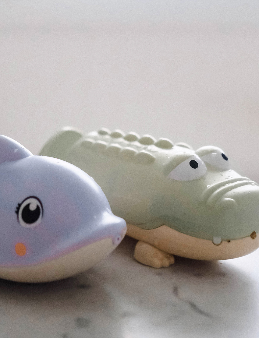 A close-up image of two ceramic water squirters on a marble surface. On the left is a purple dolphin with a smiling expression, and on the right is a light green alligator featuring cartoonish characteristics. The background is softly blurred. These are products from the Sunnylife brand.