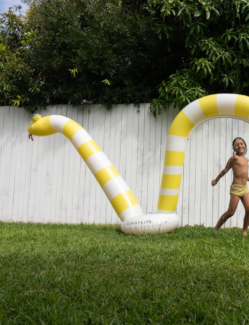 An image of the Snake Giant Sprinkler Into The Wild - Yellow by Sunnylife, an inflatable pool float crafted to resemble a snake. It features a yellow and white striped body with large eyes on its head and a small tongue sticking out. The float is designed to create three arches, with the tail forming a small loop at the end.