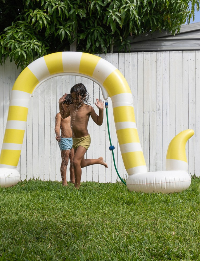 An image of the Snake Giant Sprinkler Into The Wild - Yellow by Sunnylife, an inflatable pool float crafted to resemble a snake. It features a yellow and white striped body with large eyes on its head and a small tongue sticking out. The float is designed to create three arches, with the tail forming a small loop at the end.