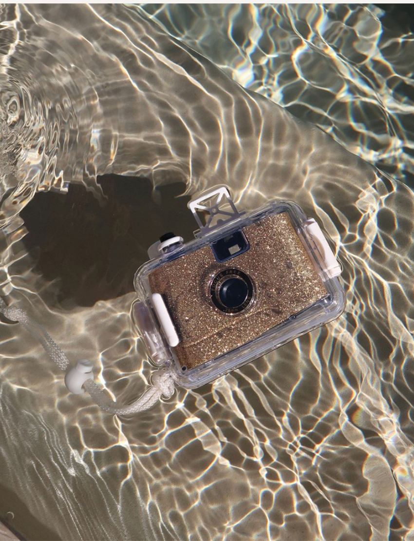 The Sunnylife Underwater Camera - Glitter Gold sits centered against a plain white background, enclosed in a transparent waterproof casing. Featuring buttons and a wrist strap, it suggests suitability for outdoor or underwater use.