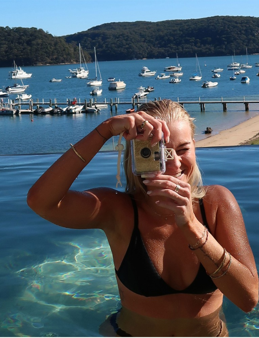 The Underwater Camera - Glitter Gold by Sunnylife floats on clear, shallow water. The sun creates rippling light patterns on the water's surface. A white cord is attached to the camera, trailing in the water.