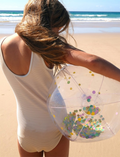 A child at the beach holds a Sunnylife Inflatable Beach Ball - Confetti Multi in front of their face. The sandy shore and ocean waves are visible in the background under a clear blue sky.