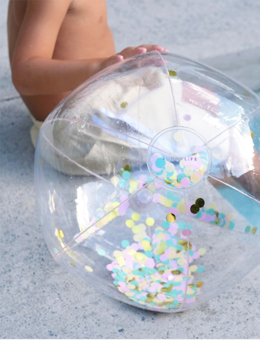 A child at the beach holds a Sunnylife Inflatable Beach Ball - Confetti Multi in front of their face. The sandy shore and ocean waves are visible in the background under a clear blue sky.