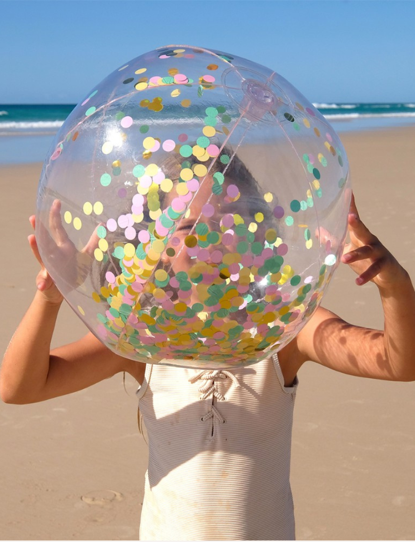 A child at the beach holds a Sunnylife Inflatable Beach Ball - Confetti Multi in front of their face. The sandy shore and ocean waves are visible in the background under a clear blue sky.