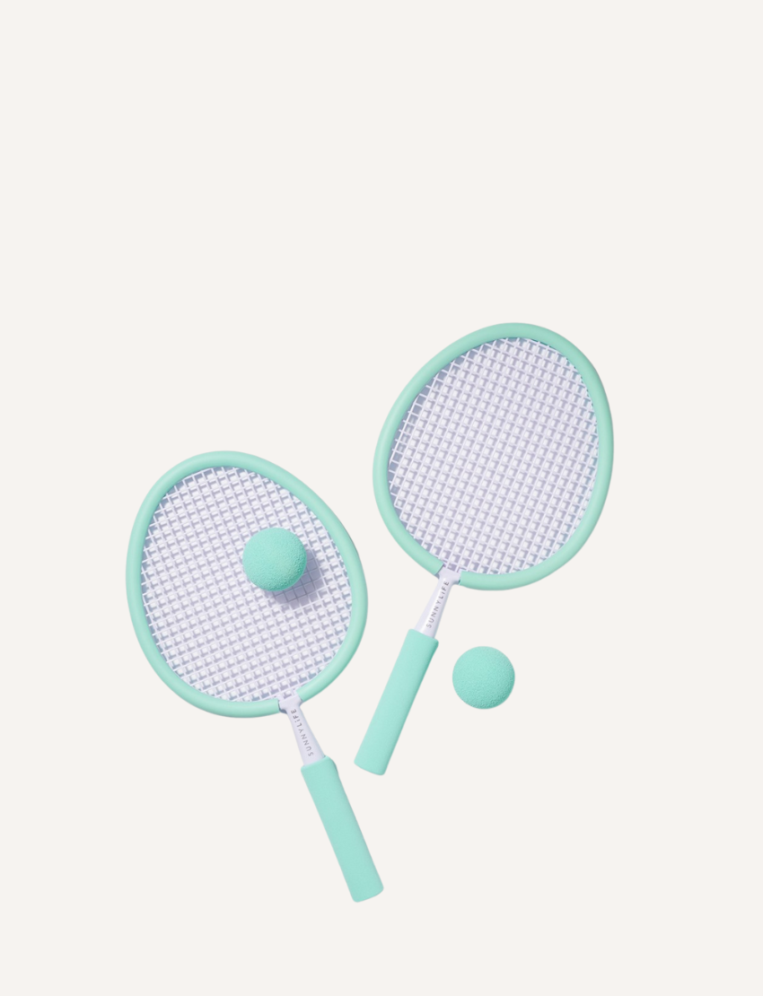 Two Sunnylife Foam Beach Bats with white netting and handles are resting against a white wall on sandy ground. Two matching light blue foam balls are placed near the rackets, suggesting a casual beach game setup.