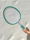 Two Sunnylife Foam Beach Bats with white netting and handles are resting against a white wall on sandy ground. Two matching light blue foam balls are placed near the rackets, suggesting a casual beach game setup.