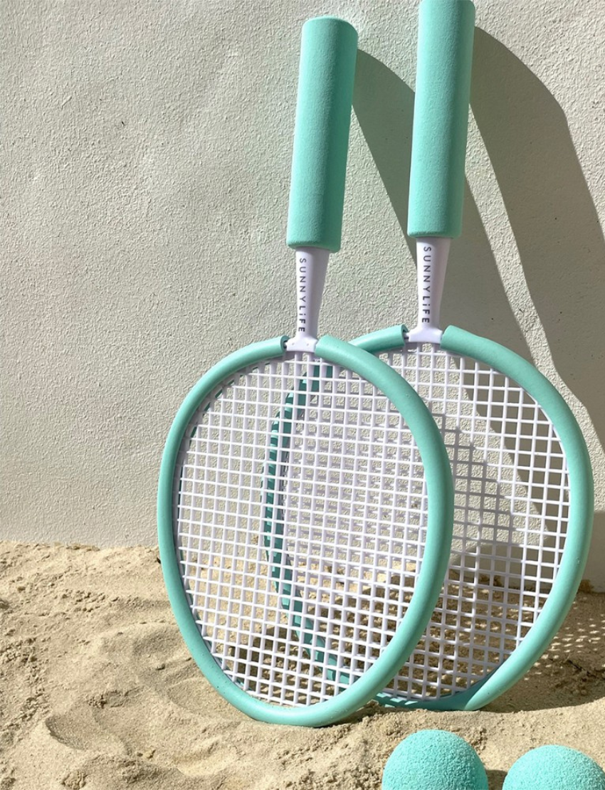 Two Sunnylife Foam Beach Bats with white netting and handles are resting against a white wall on sandy ground. Two matching light blue foam balls are placed near the rackets, suggesting a casual beach game setup.