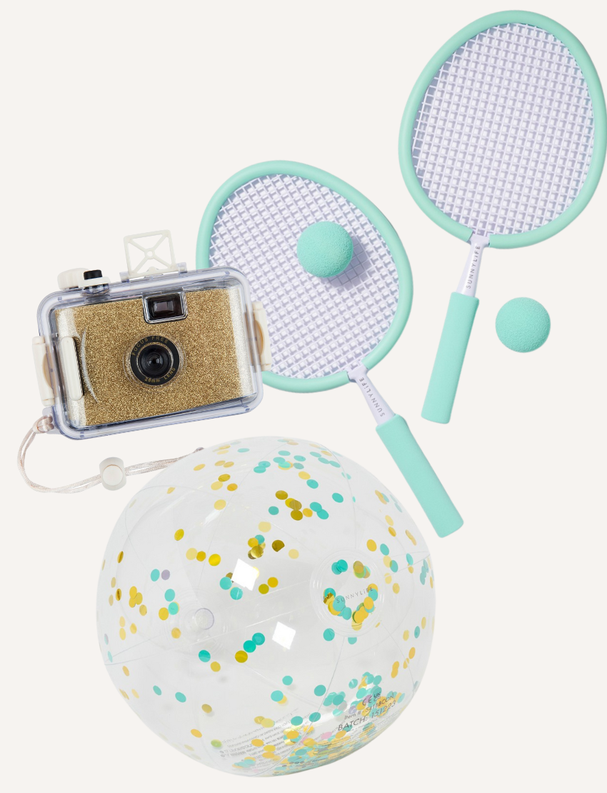 A flat lay image featuring the SunnyLife Mini Bundle by Sunnylife, including a transparent toy camera filled with glitter, two mint green badminton rackets with balls, and a clear inflatable beach ball brimming with colorful confetti.